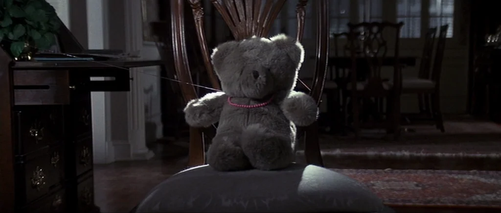 Cape Fear (1991) movie still: closeup — A dimly lit room contains a wooden chair holding a gray plush teddy bear. A thin thread attaches the bear to…