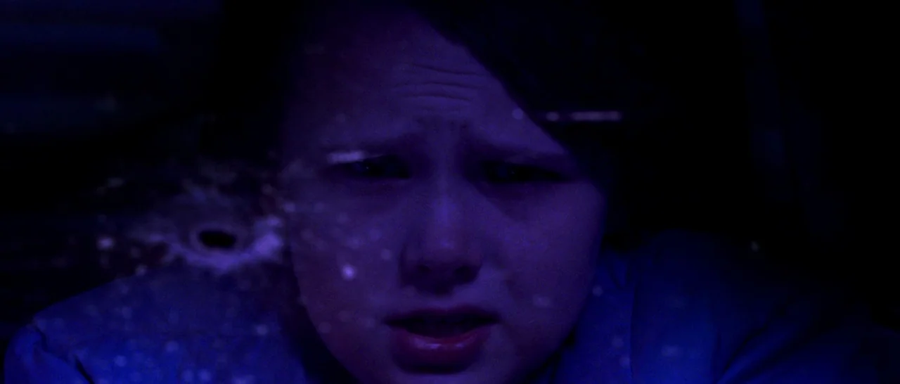 Cop Car (2015) movie still: closeup — A child sits inside a vehicle at night with blue light illuminating their face. A spiderweb-like crack in the…