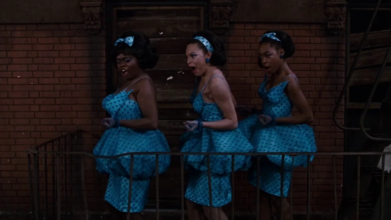 Little Shop of Horrors (1986) movie still: medium — Three young women stand on a dark outdoor metal fire escape against a brick building. They wear…