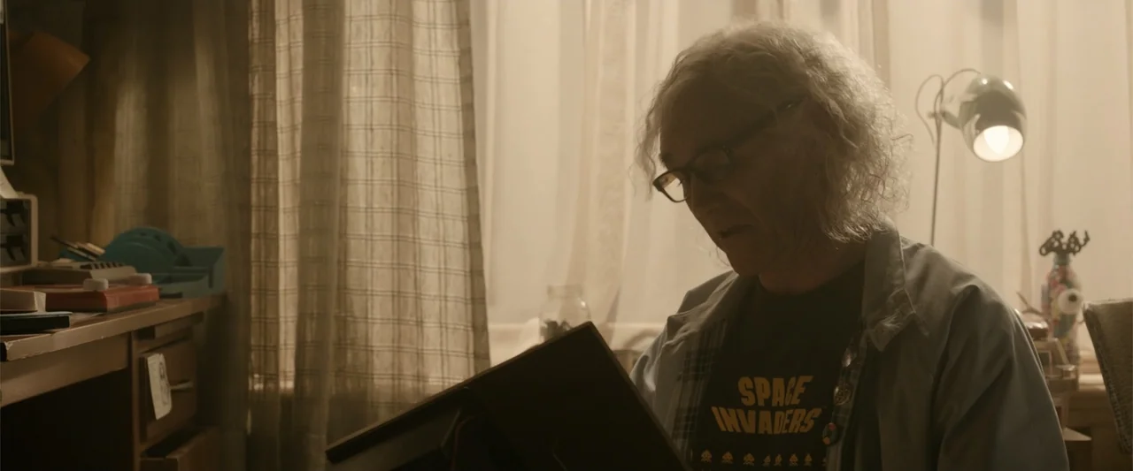 Ready Player One (2018) movie still: medium — An interior living room with sheer curtains covering a window and desk clutter visible. An elderly man…