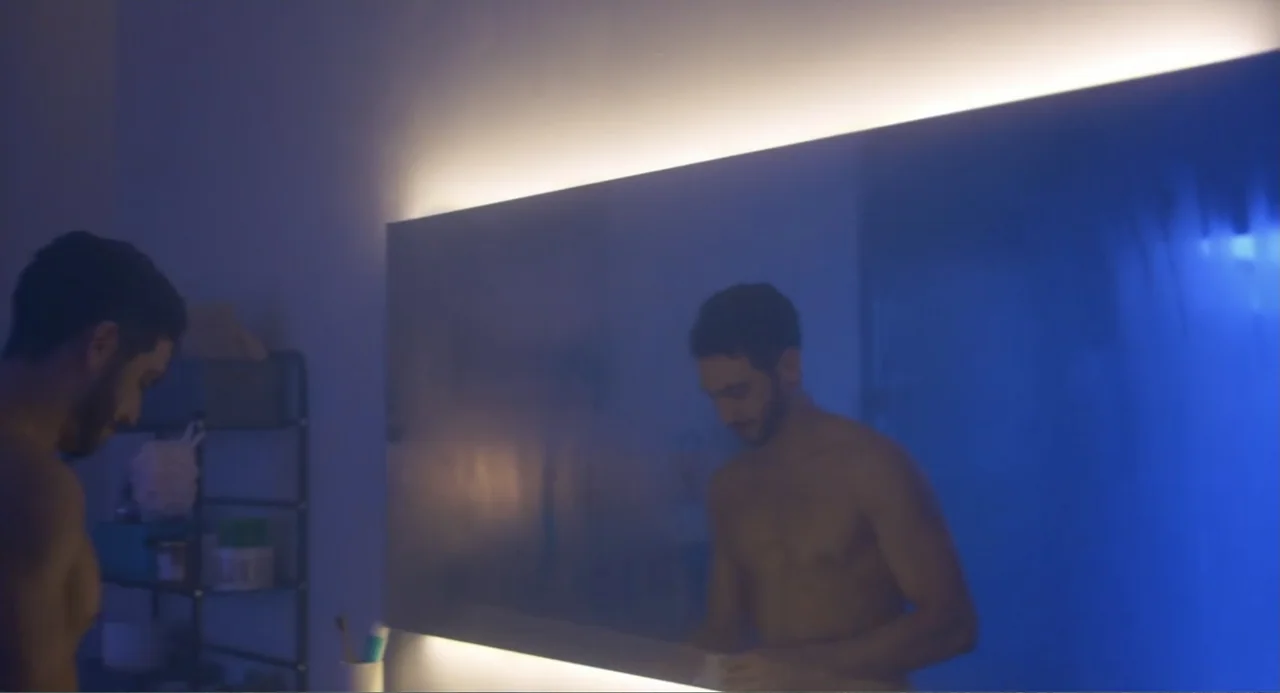 Straight (2024) movie still: medium — A modern minimalist bathroom is illuminated by a singular long horizontal light strip. A shirtless man with a…