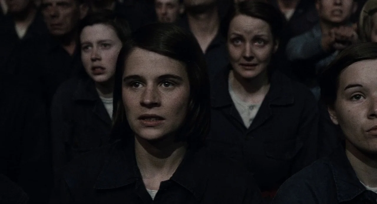 1984 (1984) movie still: medium — A group of people are gathered in a dark, poorly lit indoor space. The people wear simple, matching dark utility…