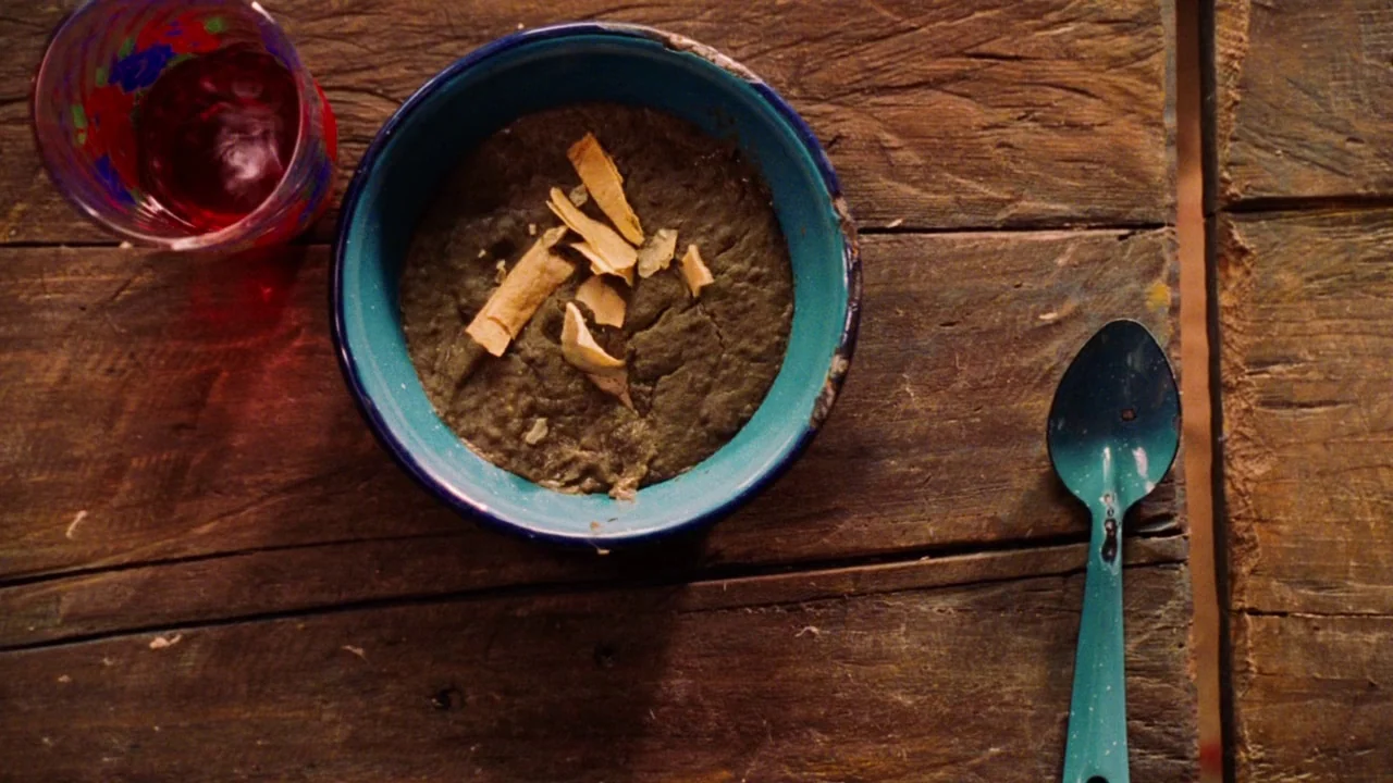 Nacho Libre (2006) movie still: closeup — A dark blue enamel bowl filled with brown stew sits on a rustic wooden table. A red glass and a matching…