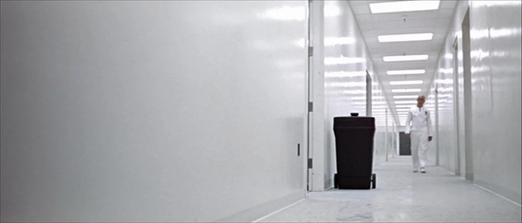 THX 1138 (1971) movie still: wide — A long, brightly lit, sterile hallway features smooth white walls and a white floor. A bald man in white…