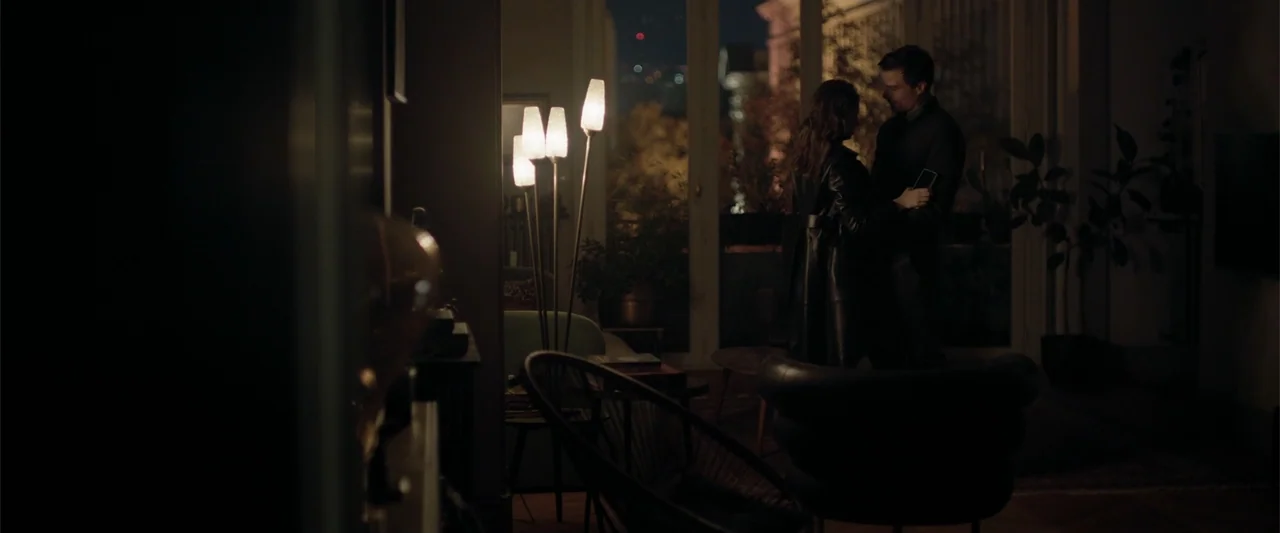 Marked Men: Rule + Shaw (2025) movie still: wide — A dimly lit room at night features a tall floor lamp casting warm light. A man and a woman stand…