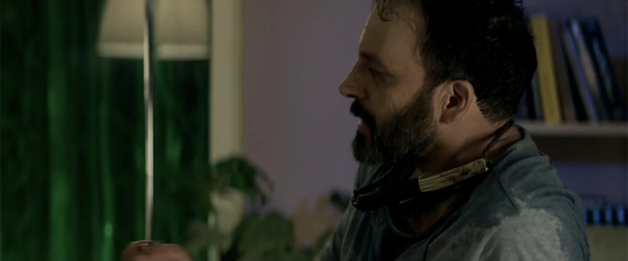Human Animals (2020) movie still: medium — A dimly lit room features a blurred bookshelf and green curtains in the back. A man with a metal…