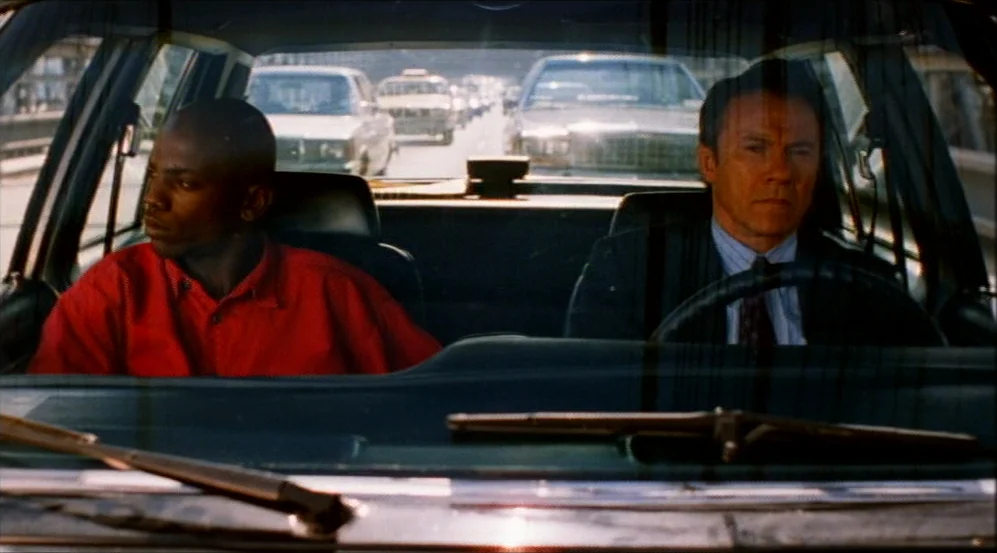 Clockers (1995) movie still: medium — Two people sit inside a car parked amidst a line of traffic on a bridge. The man drives while the boy sits in…