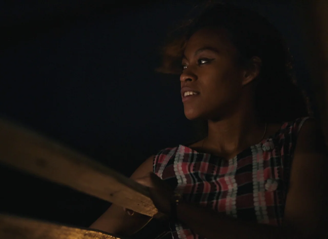 Red Island (2023) movie still: medium — A dark night setting features a woman standing at a wooden outdoor railing. The background is completely…