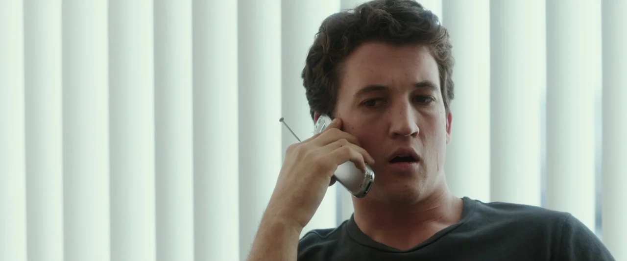 War Dogs (2016) movie still: closeup — A man stands before vertical white window blinds in an interior space. The subject holds a silver mobile…