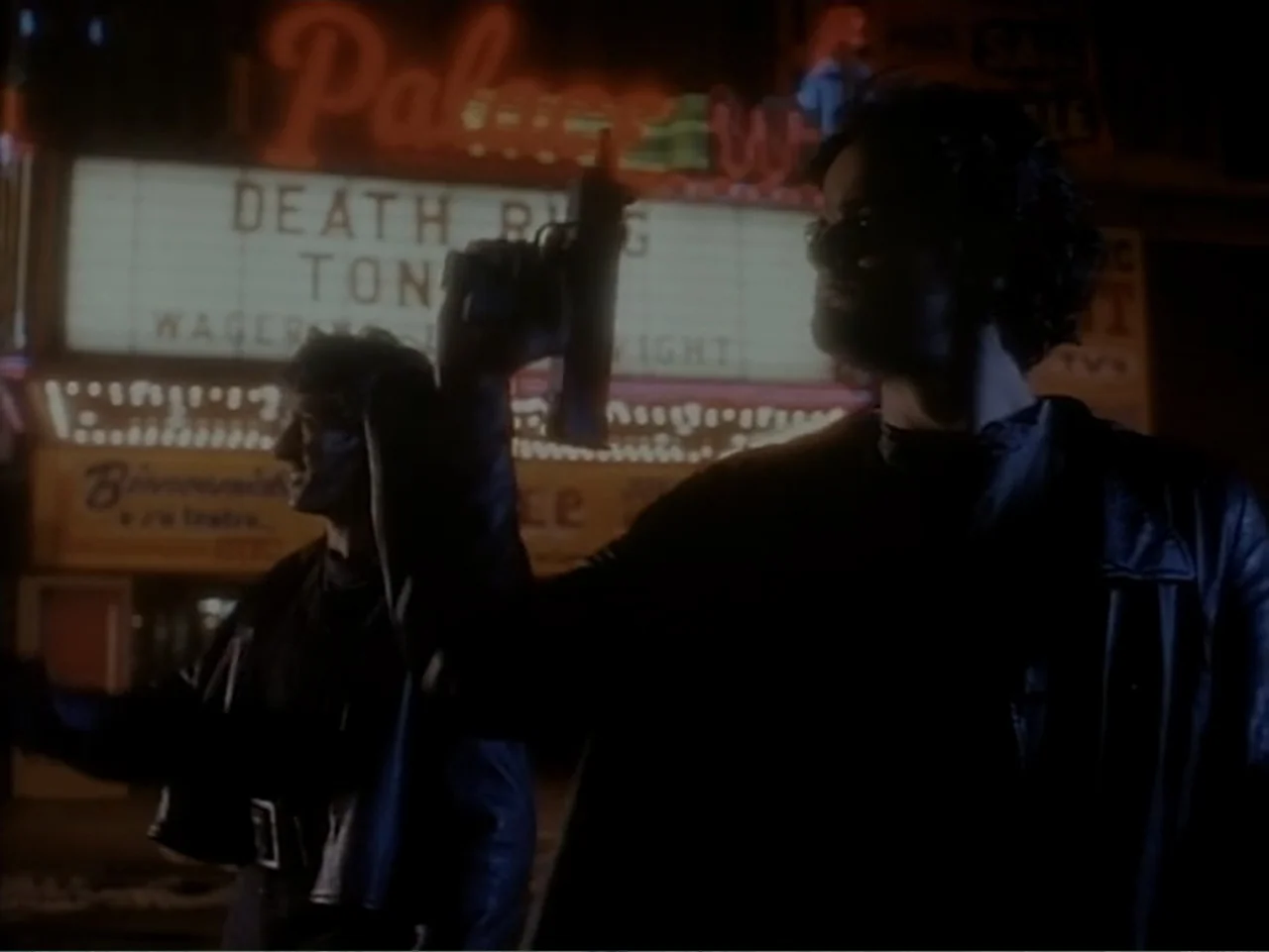 Firepower (1979) movie still: medium — A dark city sidewalk at night in front of a brightly lit movie theater marquee. Two men in leather jackets…
