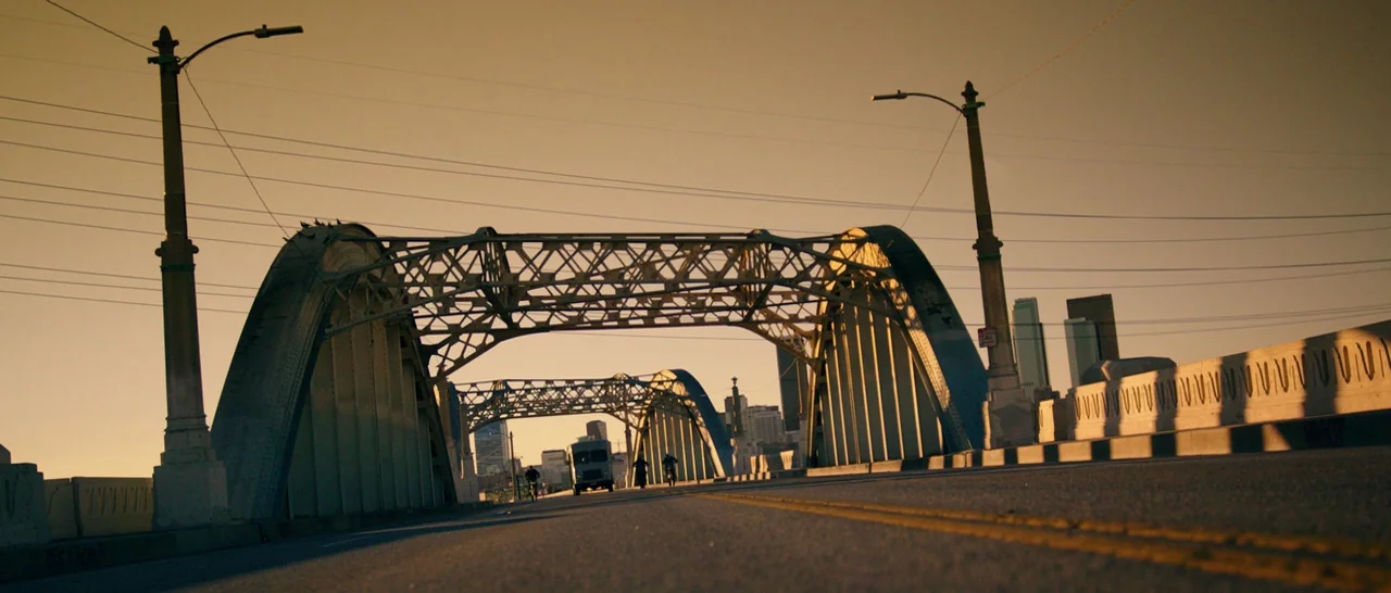 The Purge: Anarchy (2014) movie still: establishing — A low-angle view of a historic steel arch bridge with concrete pillars at sunset. The…