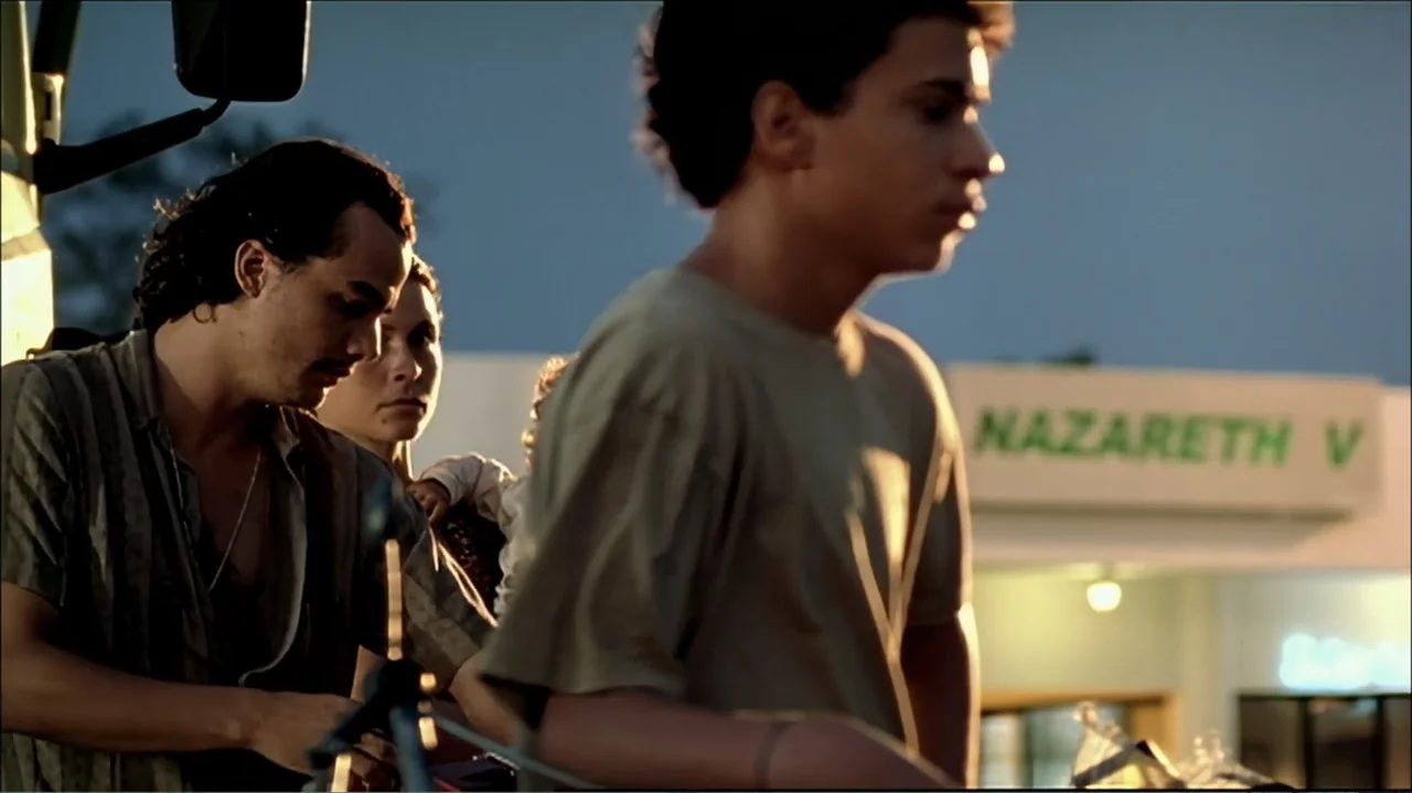 In the Middle of the World (1990) movie still: medium — A gas station exterior features a sign reading Nazareth V under golden hour light. Three…