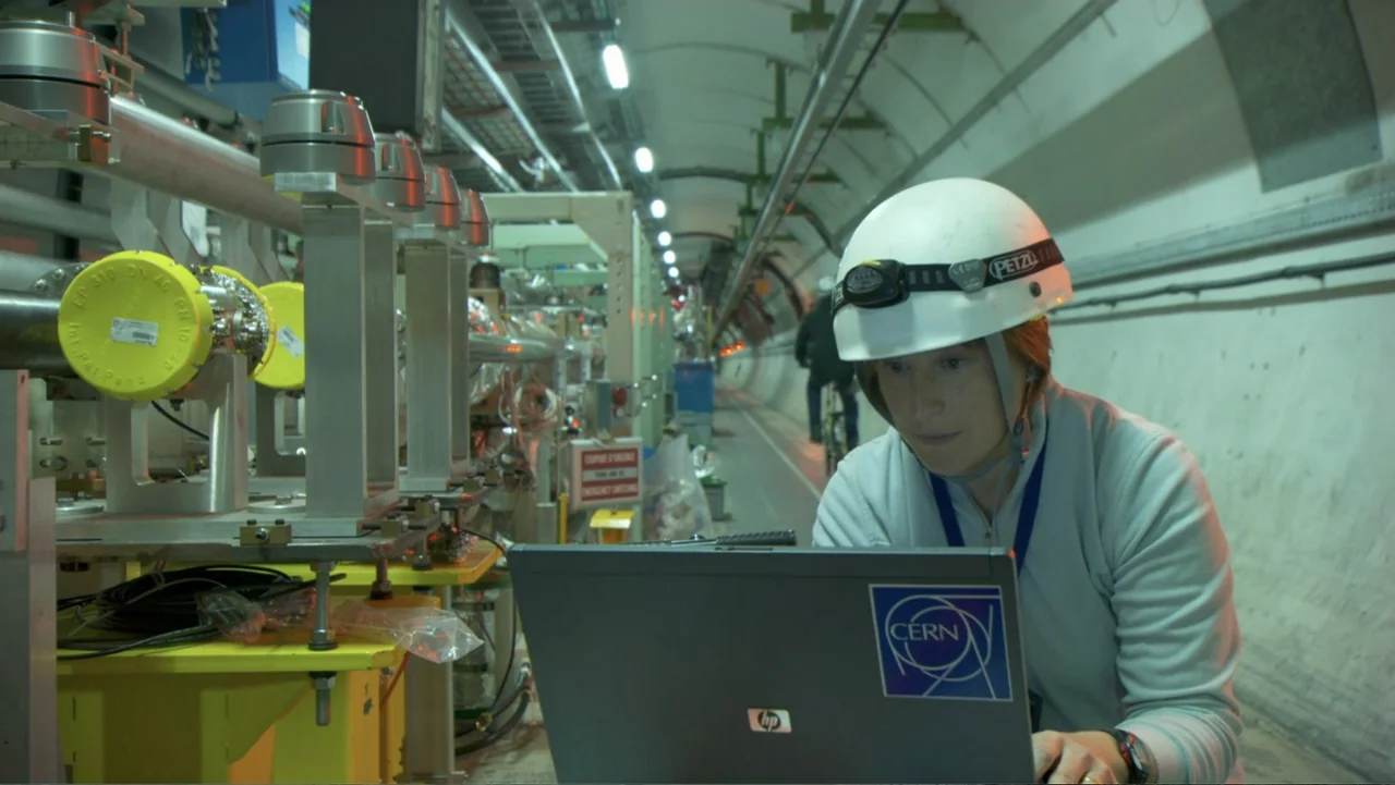 CERN (2013) movie still: medium — A long, curved concrete tunnel houses complex scientific machinery and metallic piping. A woman in a hard hat sits…