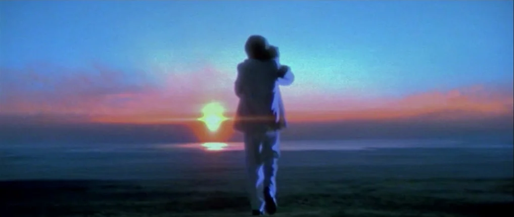 Titus (1999) movie still: fullbody — A figure in a light-colored suit stands on a shoreline facing the horizon. The sun is setting over the water in…
