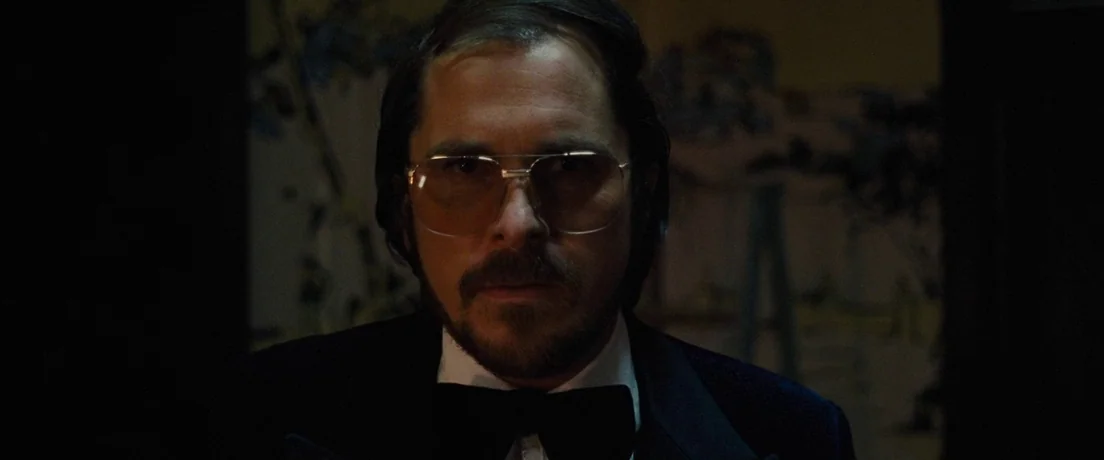 American Hustle (2013) movie still: closeup — A dimly lit event space with dark surroundings and blurred architectural shapes behind. A man with a…