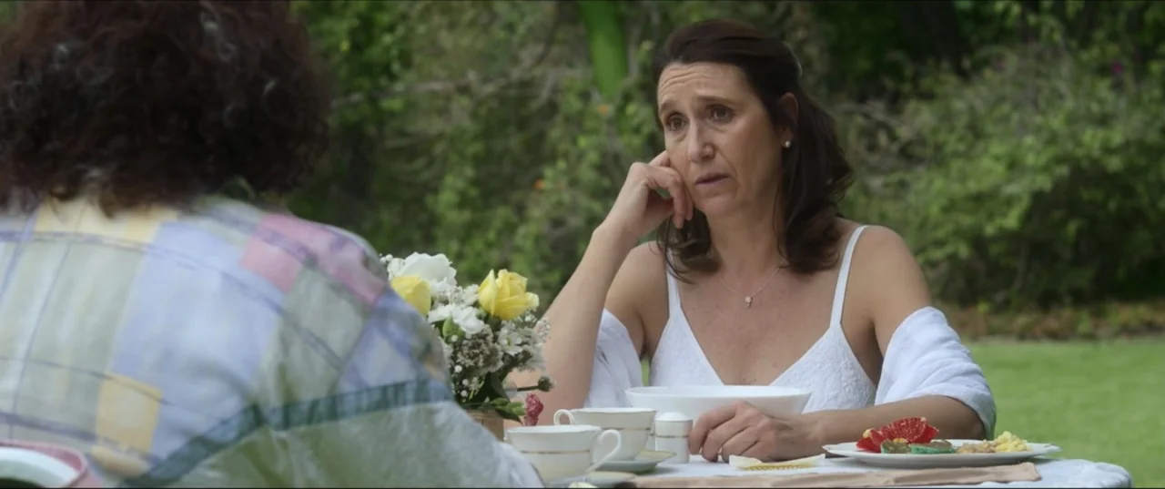 Can't Quit Mom (2022) movie still: medium — A woman sits at an outdoor table with a white tablecloth and floral centerpiece. She listens while…