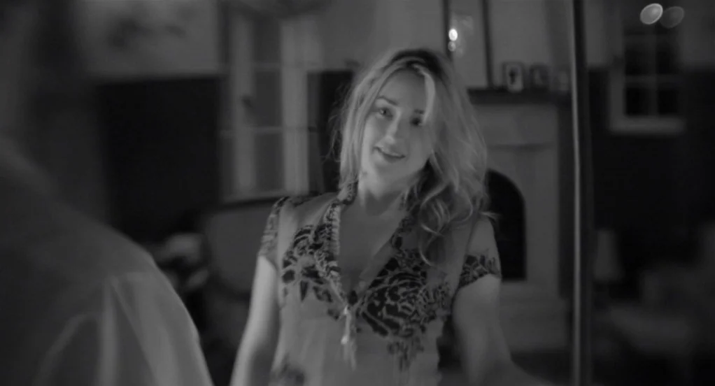Much Ado About Nothing (2012) movie still: medium — A dimly lit room with a fireplace and furniture in the background. A woman in a floral top…