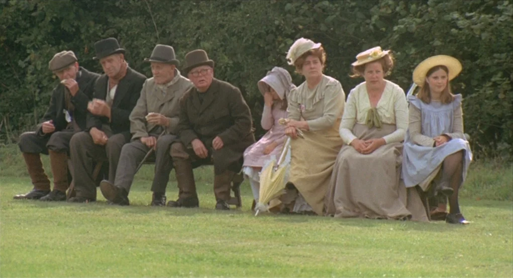 The Go-Between (1971) movie still: wide — A group of men and women sit on a long bench in a green field. They watch an off-screen event while…
