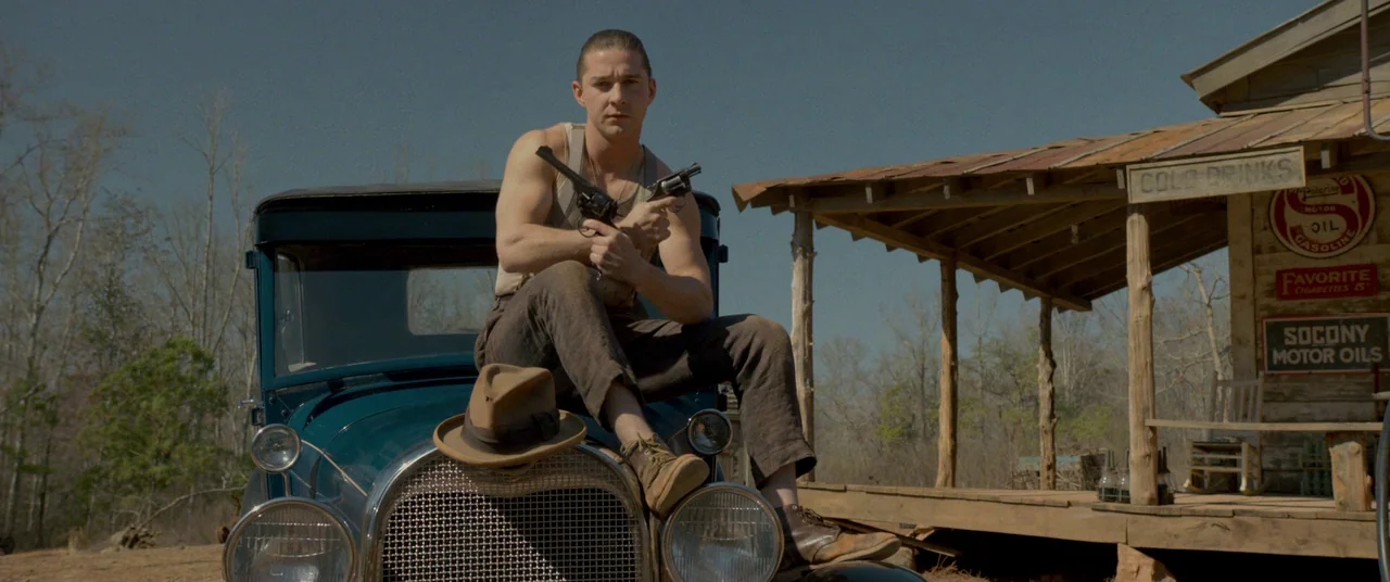 Lawless (2012) movie still: medium — A rustic wood building with advertising signs stands next to an old blue automobile. A man in period work…