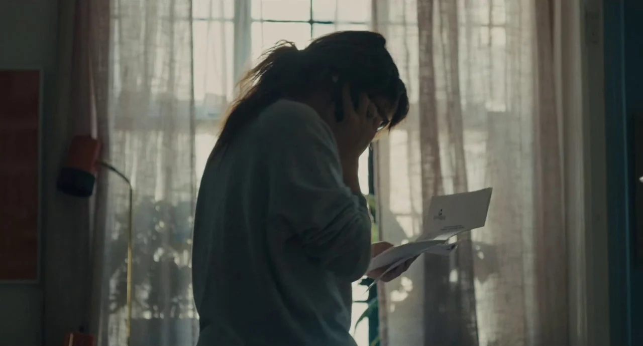 Private Life (2018) movie still: medium — A woman stands in silhouette against a brightly lit window with sheer curtains. She holds a document in…