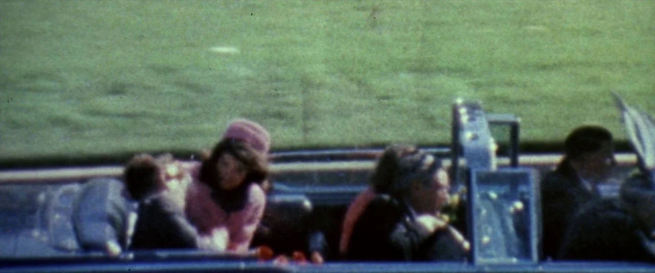 JFK (1991) movie still: medium — A group of people sit in the open rear seat of a moving limousine. The car travels along a bright green grass verge…