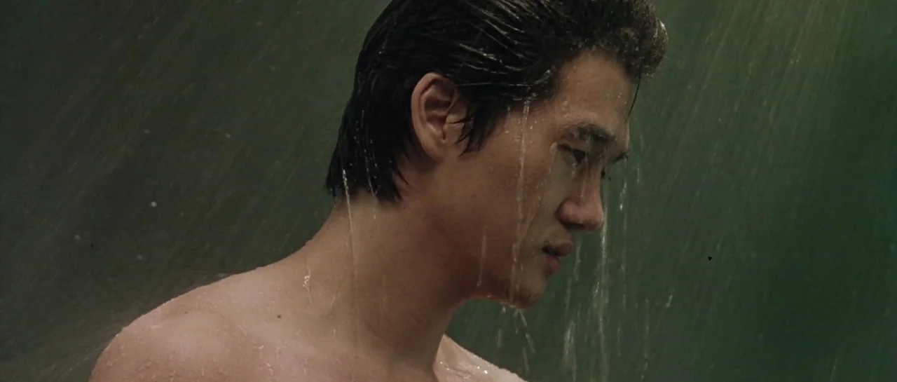 Oldboy (2003) movie still: closeup — A man stands beneath a continuous stream of water within an enclosed space. Water drips from his slicked hair…