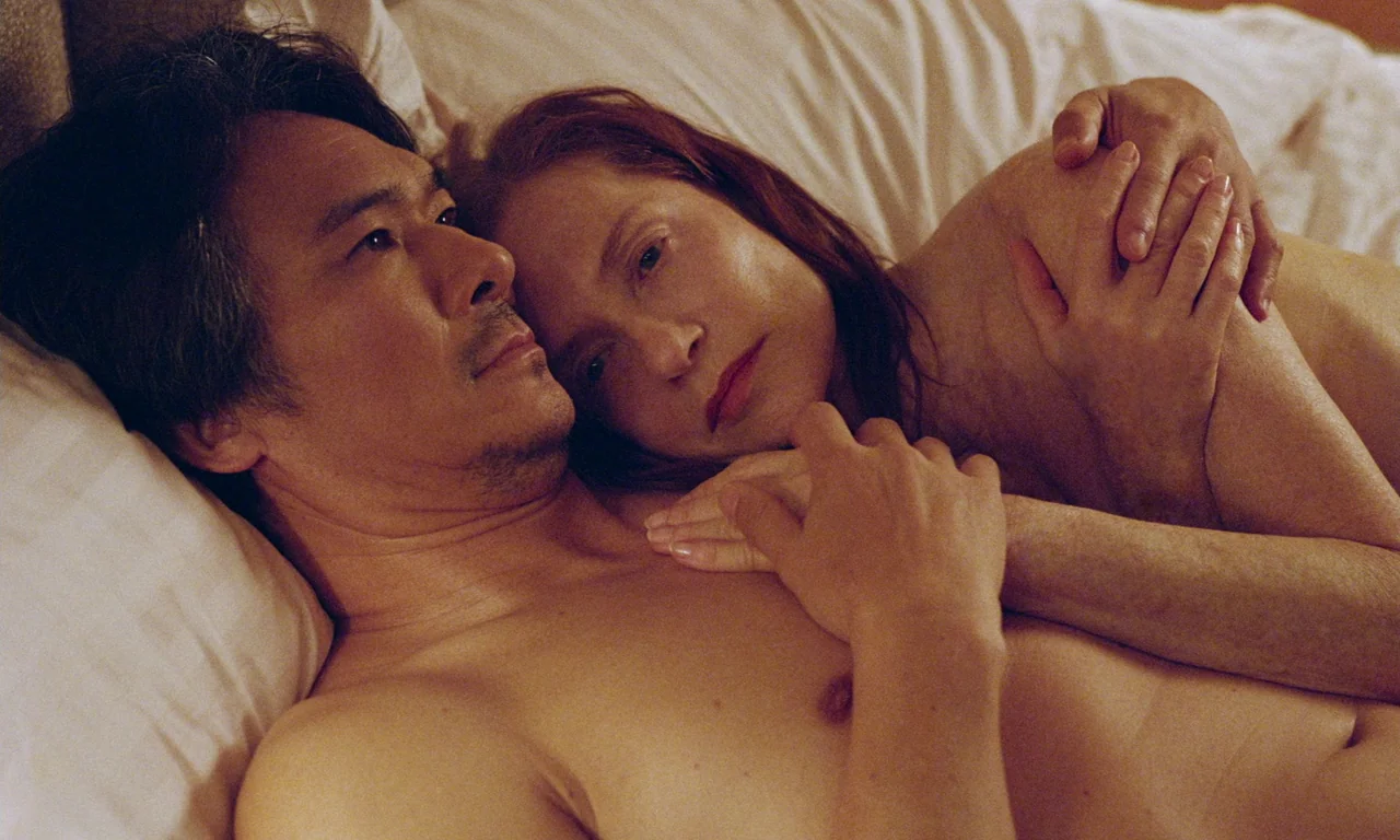 Sidonie in Japan (2023) movie still: closeup — A man and a woman lie in a bed with rumpled white linens. They are resting close together, with their…