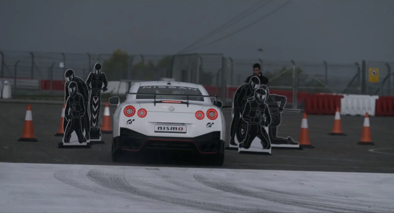 Gran Turismo (2023) movie still: medium — A white sports car drives away on a paved track marked by orange cones. Cardboard cutouts of people are…