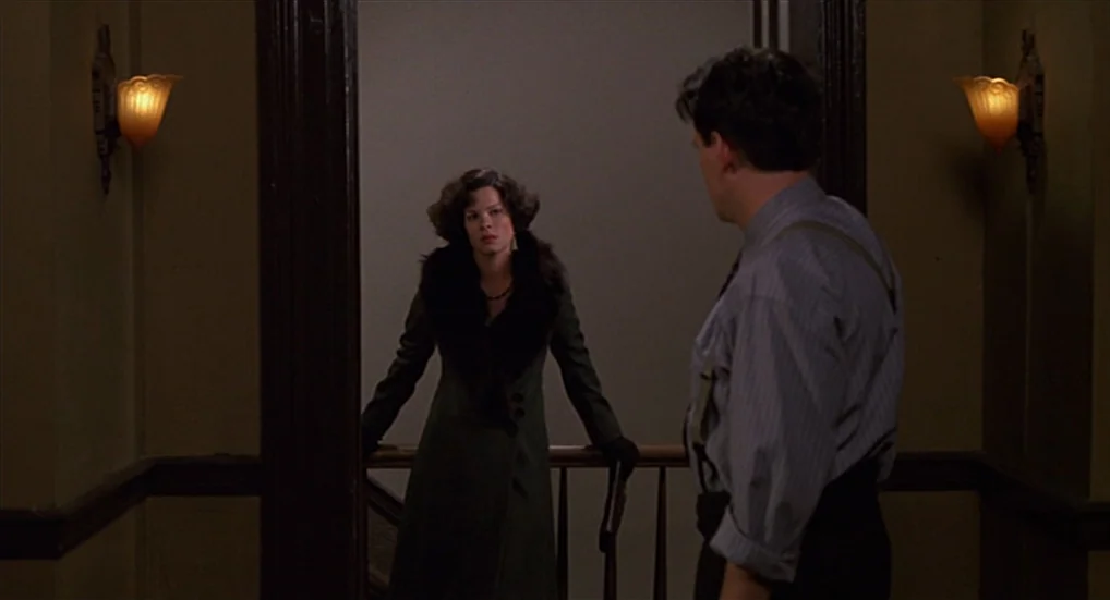Miller’s Crossing (1990) movie still: medium — A dimly lit indoor landing features dark wooden trim and amber wall sconces. A woman in a fur-trimmed…