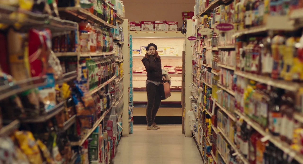 Mistress America (2015) movie still: wide — A long, narrow aisle is lined with high shelves packed with colorful grocery products. A woman in a…