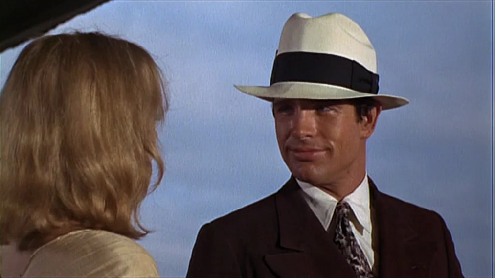 Bonnie And Clyde (1967) movie still: medium — A man and woman stand together outdoors against a clear blue sky background. The man wears a vintage…