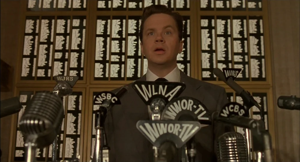 The Hudsucker Proxy (1994) movie still: medium — A man stands in front of a grid of small white name cards. He is speaking at a cluster of…