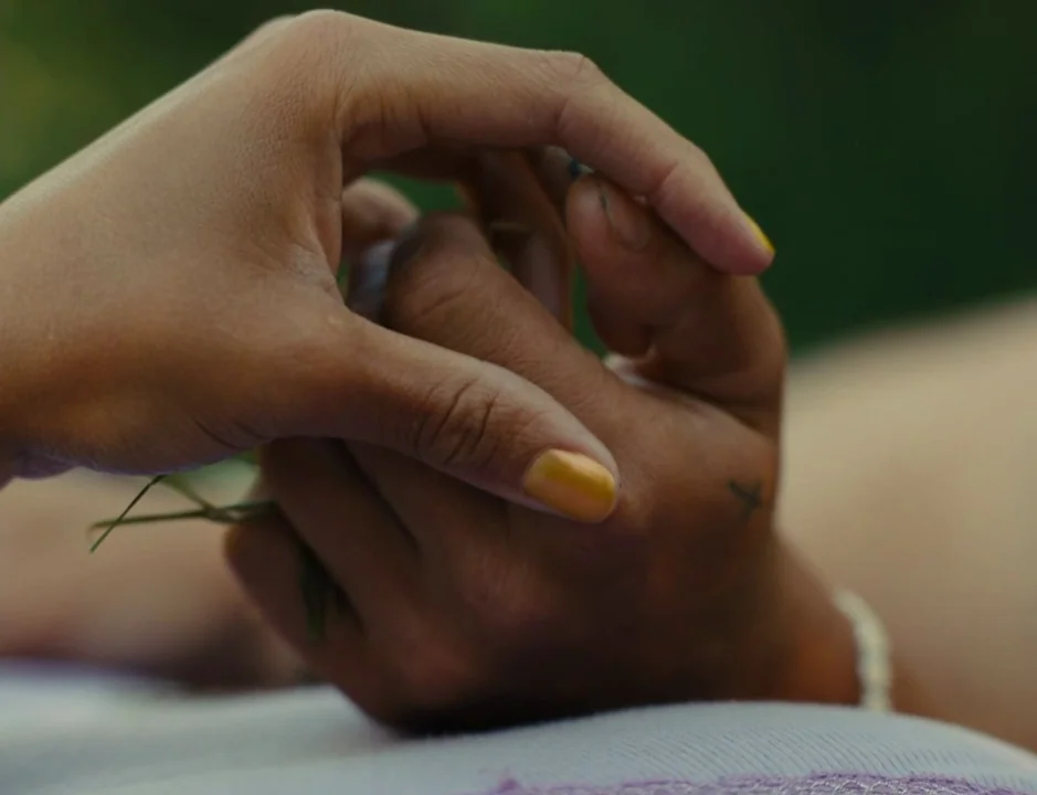 American Honey (2016) movie still: closeup — Two hands are intertwined resting against a light purple fabric surface. A small cross tattoo is…