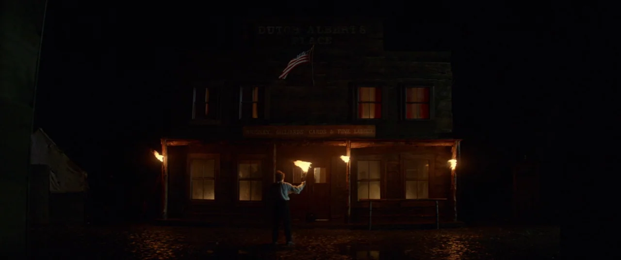 Never Grow Old (2019) movie still: wide — A dark wooden saloon building features a central flag and an overhanging porch roof. A man stands in the…