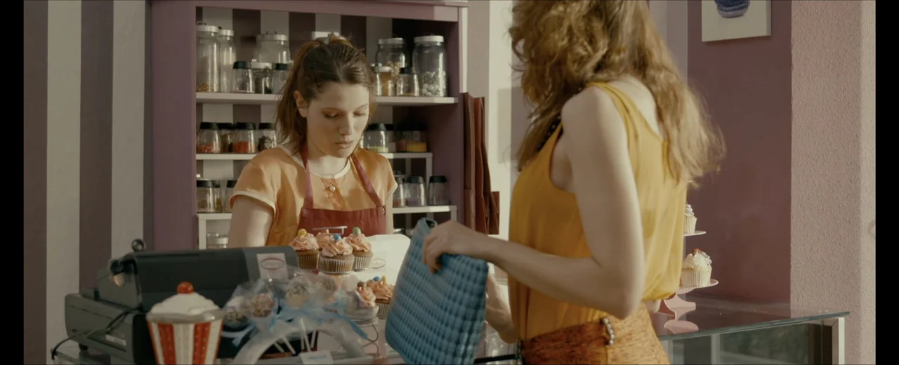 Faraday (2013) movie still: medium — A small bakery interior with pink walls and shelves displaying glass jars of sweets. A store clerk in an apron…