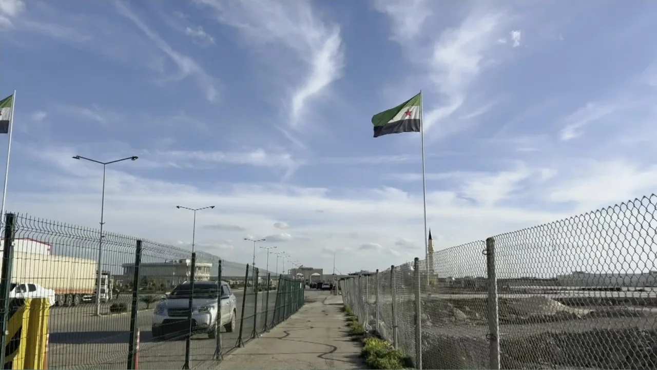 Death Without Mercy (2024) movie still: wide — A secured border crossing area featuring tall chain-link fences and a paved path. A flag with green,…
