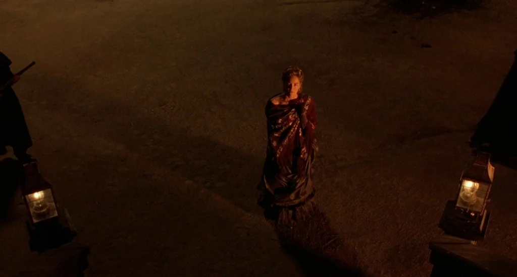 The Quick and the Dead (1995) movie still: wide — An outdoor paved area is illuminated by two glowing lanterns at the bottom edge. A woman stands…