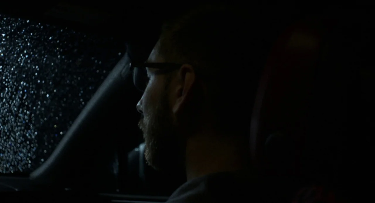 Happer’s Comet (2022) movie still: closeup — A man sits in the driver's seat of a car at night during rain. Raindrops cover the windshield, blurring…