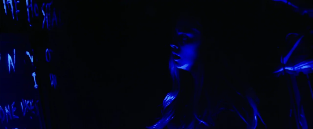 Lights Out (2016) movie still: closeup — A dimly lit interior space features blue neon writing on a dark wall. A young woman stands looking toward…