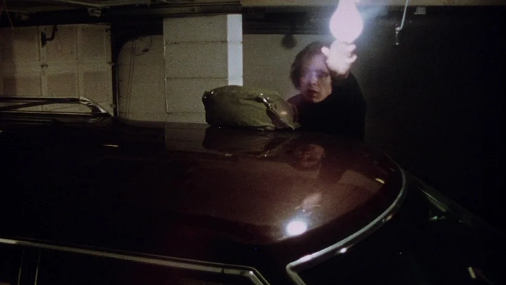 Martin (1977) movie still: medium — A dark, cramped residential space with a concrete pillar and low ceiling. A man stands near a car, extending his…