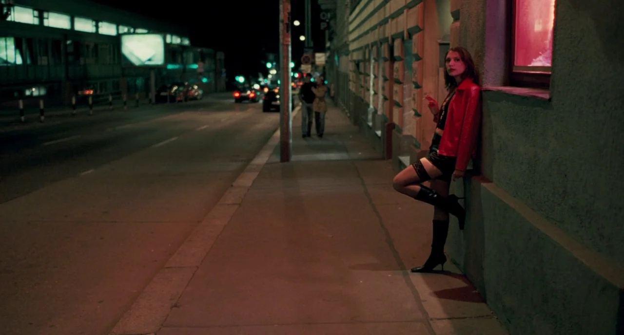 Revanche (2008) movie still: fullbody — A dark urban sidewalk at night features a textured wall under red neon light. A woman leans against the wall…