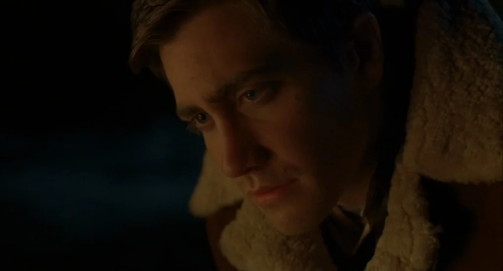 Brokeback Mountain (2005) movie still: closeup — A man sits outdoors at night by a campfire. He gazes downward while wearing a sherpa-lined leather…