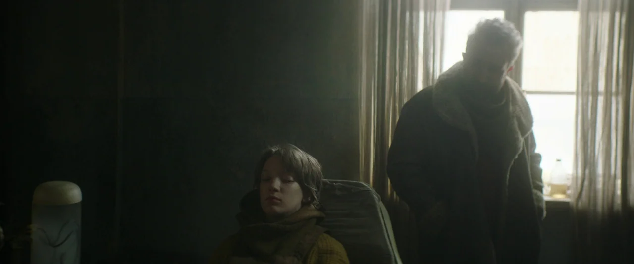 Vesper (2022) movie still: medium — A dimly lit space contains a person seated in a chair and a standing adult. The primary light source is a single…