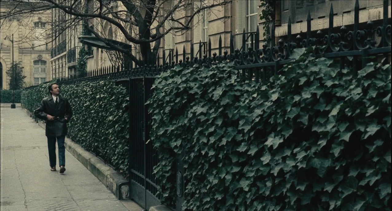 The Army of Shadows (1969) movie still: wide — A quiet sidewalk borders a tall stone building behind a dense ivy-covered metal fence. A man in a…