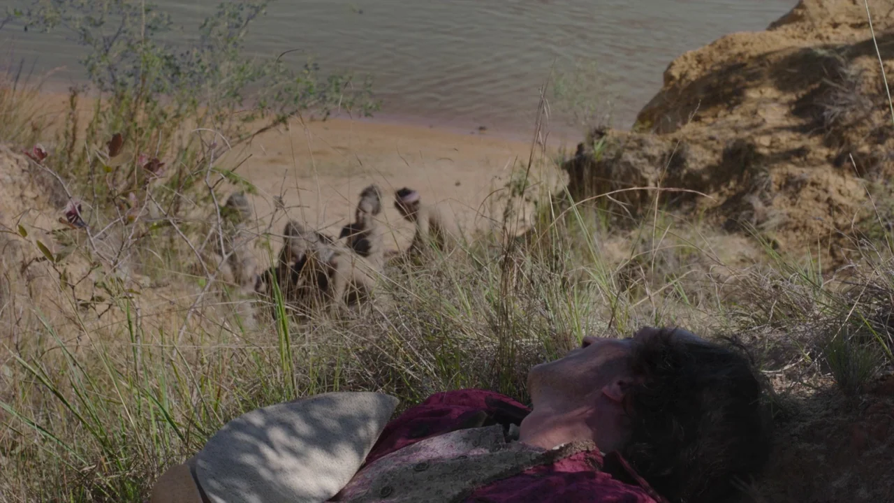 Zama (2017) movie still: medium — A man lies prone on a grassy riverbank near a flowing brown waterway. Further down the bank, several figures stand…