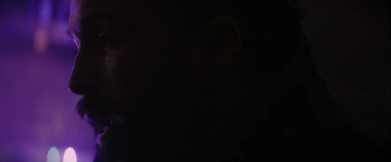 A Working Man (2025) movie still: closeup — A dimly lit interior space features deep purple lighting and heavy shadows. A man in profile looks…