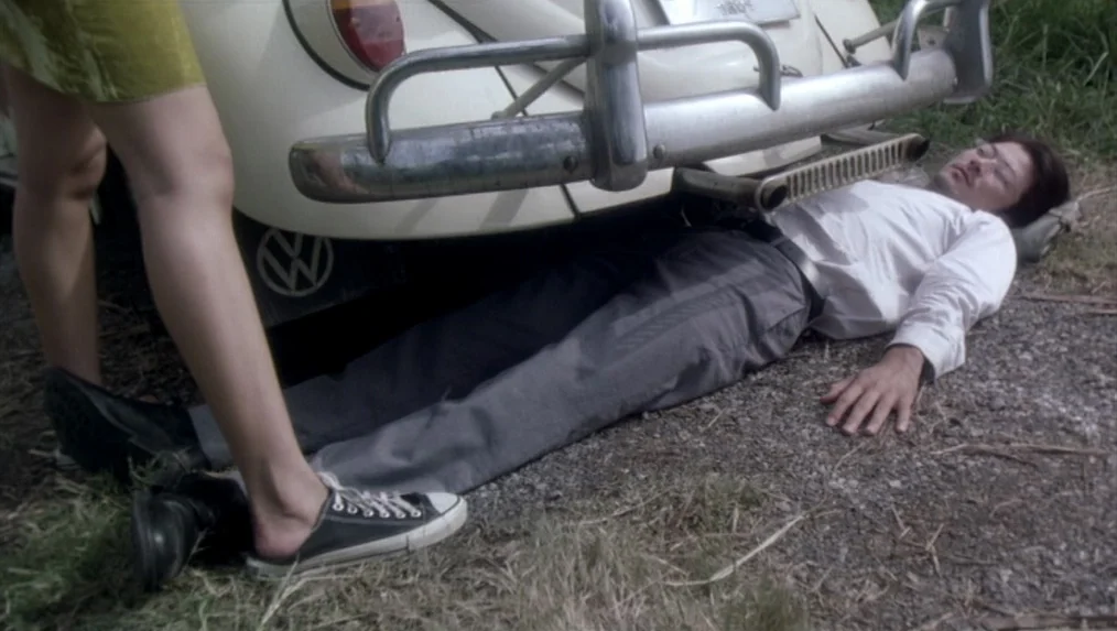 Last Life in the Universe (2003) movie still: medium — A gravel roadside with tall grass borders the rear bumper of a white vehicle. A man in a…