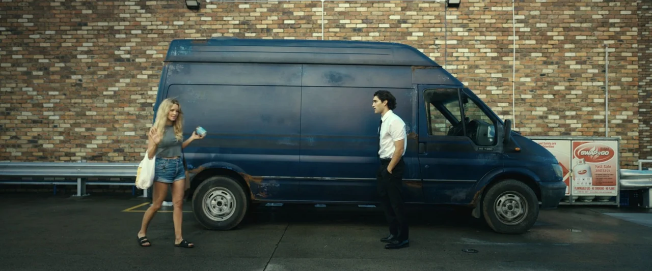 Dangerous Animals (2025) movie still: wide — A paved lot features a worn blue cargo van parked against a brick wall. A woman walks past the side of…