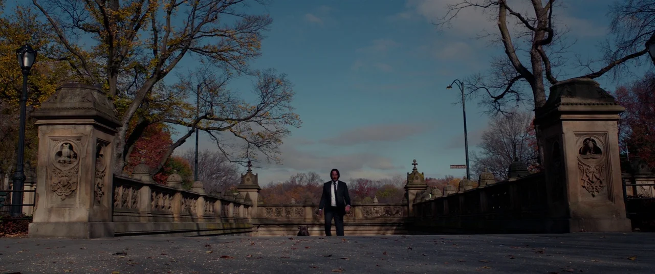 John Wick: Chapter 2 (2017) movie still: wide — A stone bridge in an urban park features ornamental pillars and autumn-leaf strewn pavement. A man…