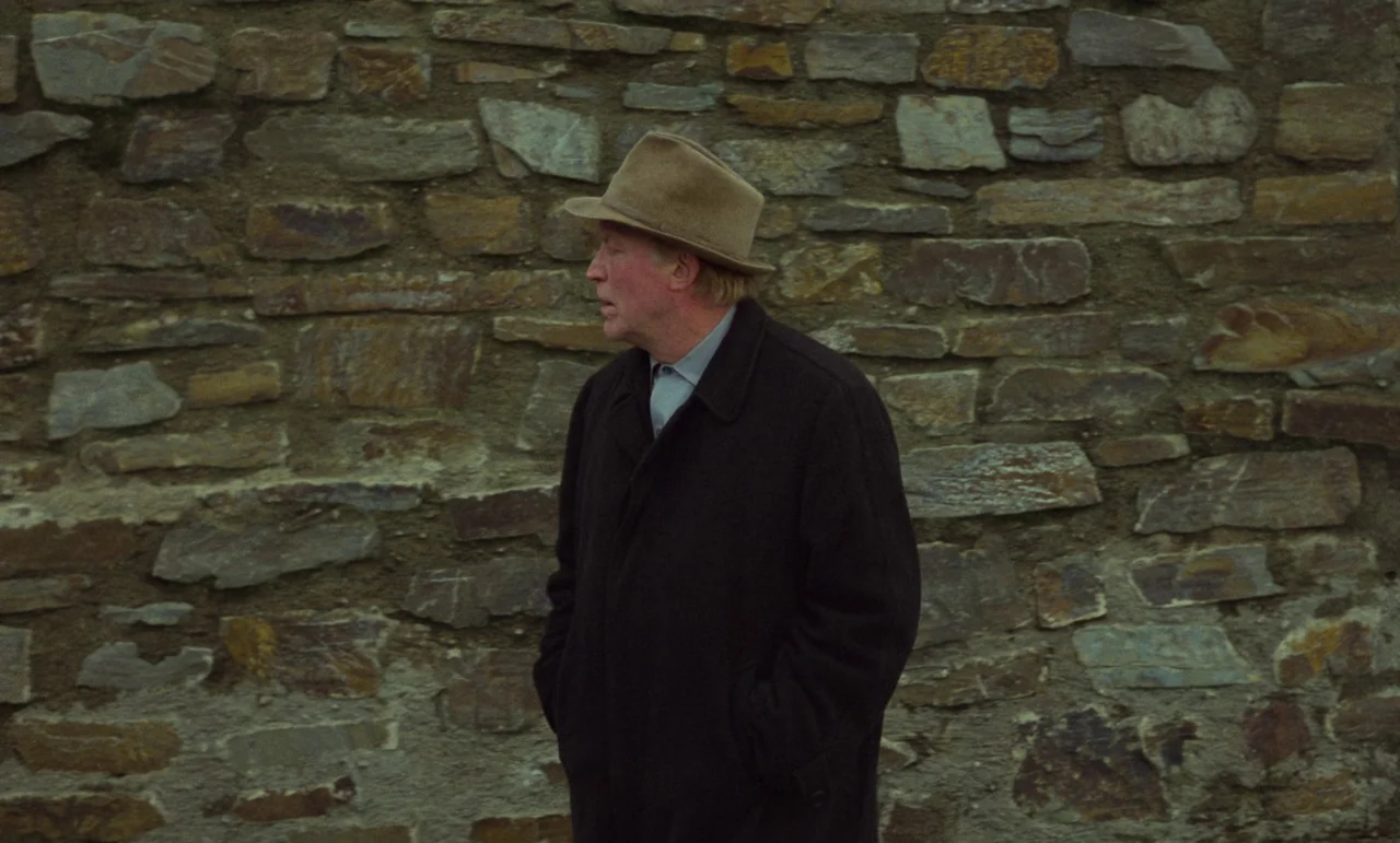 Wrong Move (1975) movie still: medium — A man stands before a rustic, mortared stone wall of varying irregular brown rocks. He wears a dark coat and…